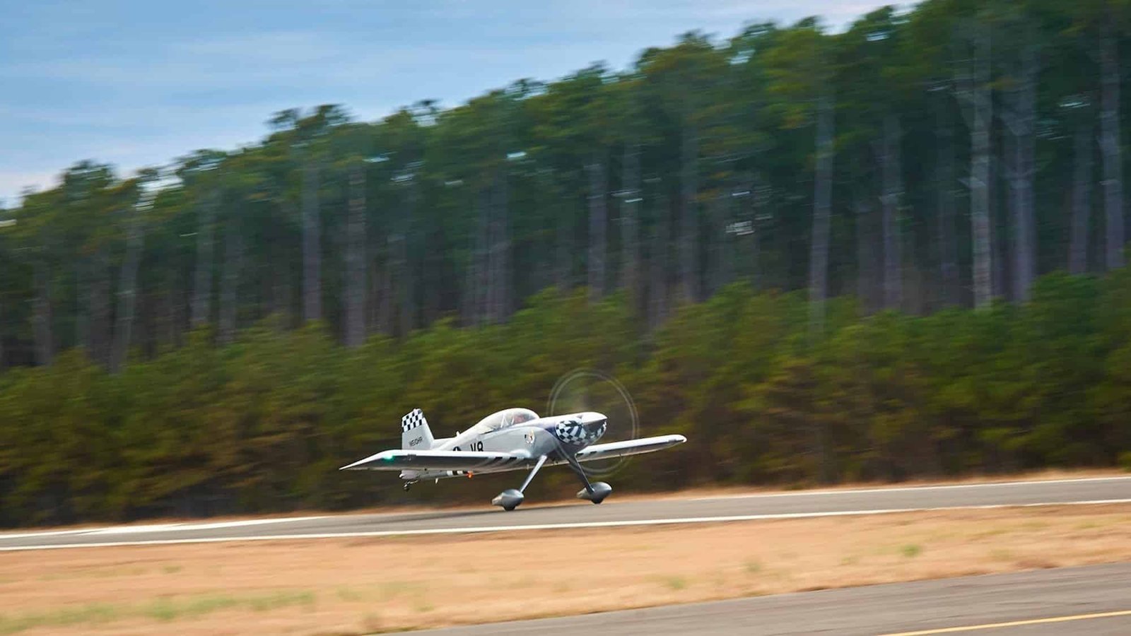 A single-engine light aircraft lifting off the 3,000-foot asphalt runway at First Flight Airport (KFFA) in Kill Devil Hills, North Carolina, with a tall pine treeline behind the field.