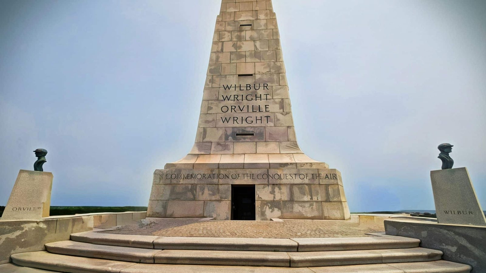 Looking up at the granite Wright Brothers Memorial pylon, carved with WILBUR WRIGHT and ORVILLE WRIGHT above the inscription IN COMMEMORATION OF THE CONQUEST OF THE AIR, with the bronze busts of the two brothers on either side.